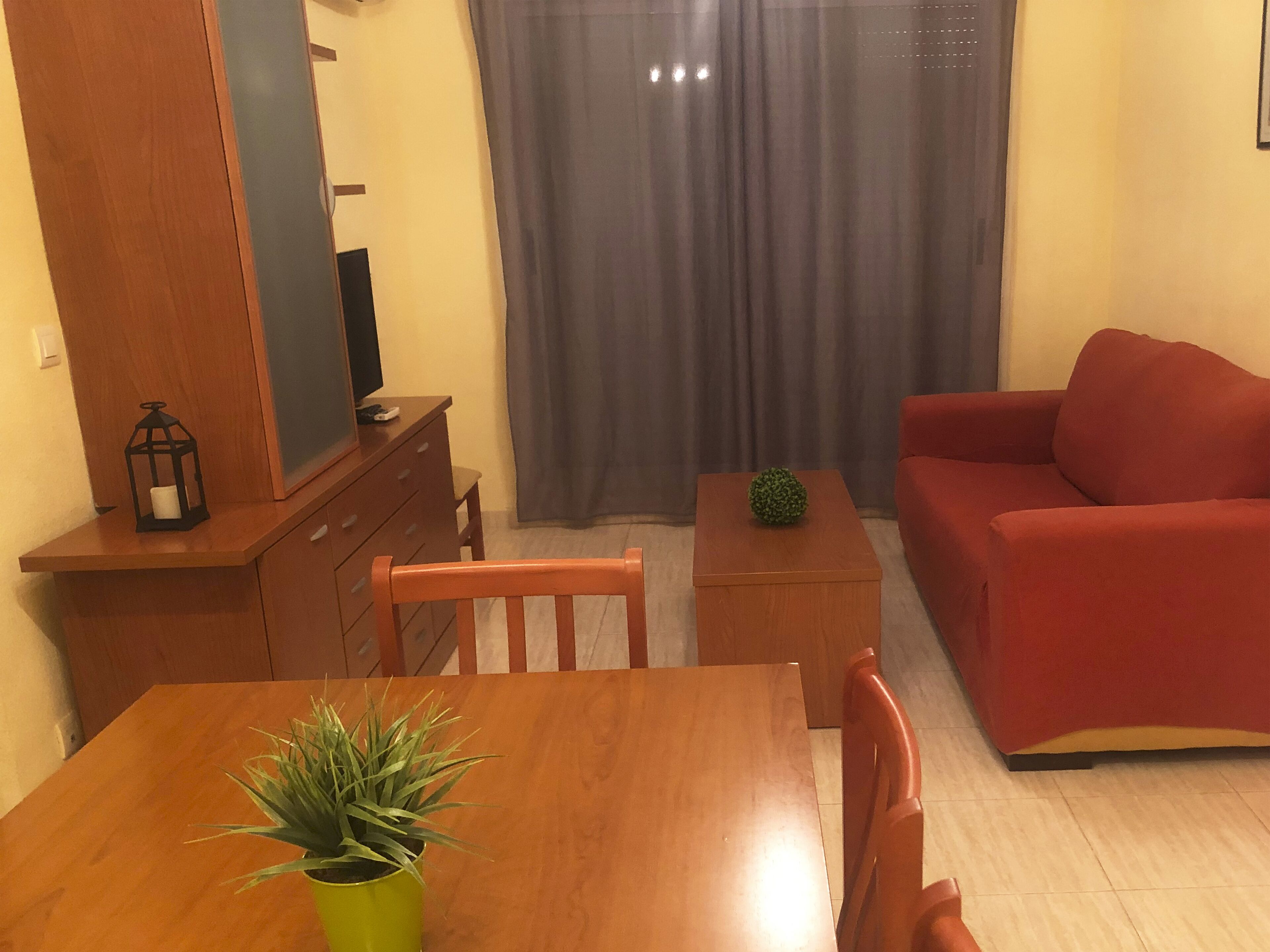 Uma sala de estar com uma mesa de jantar de madeira, um sofá vermelho, um pequeno armário de madeira e uma planta em vaso.