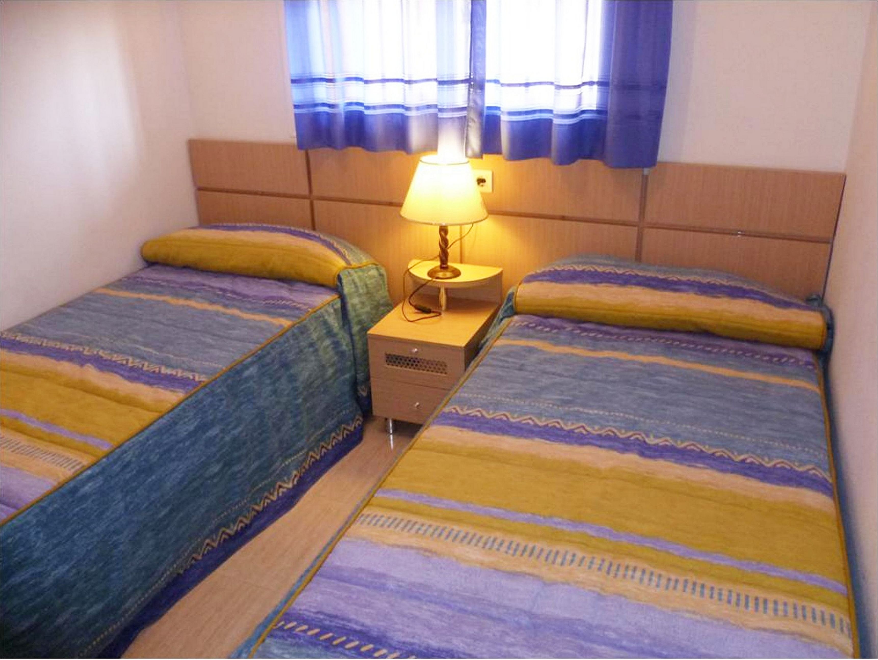 Une chambre avec deux lits, chacun recouvert d’une couvre-lit à motifs, une table de chevet avec une lampe et une fenêtre ornée de rideaux bleus.