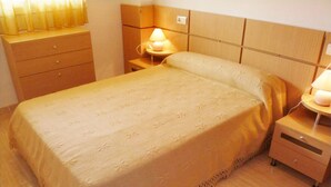 A neatly made bed with a floral-patterned bedspread, a bedside table with a lamp, and a wooden nightstand with drawers. - Apartamentos Marina D'Or 3000 (Oropesa del Mar)