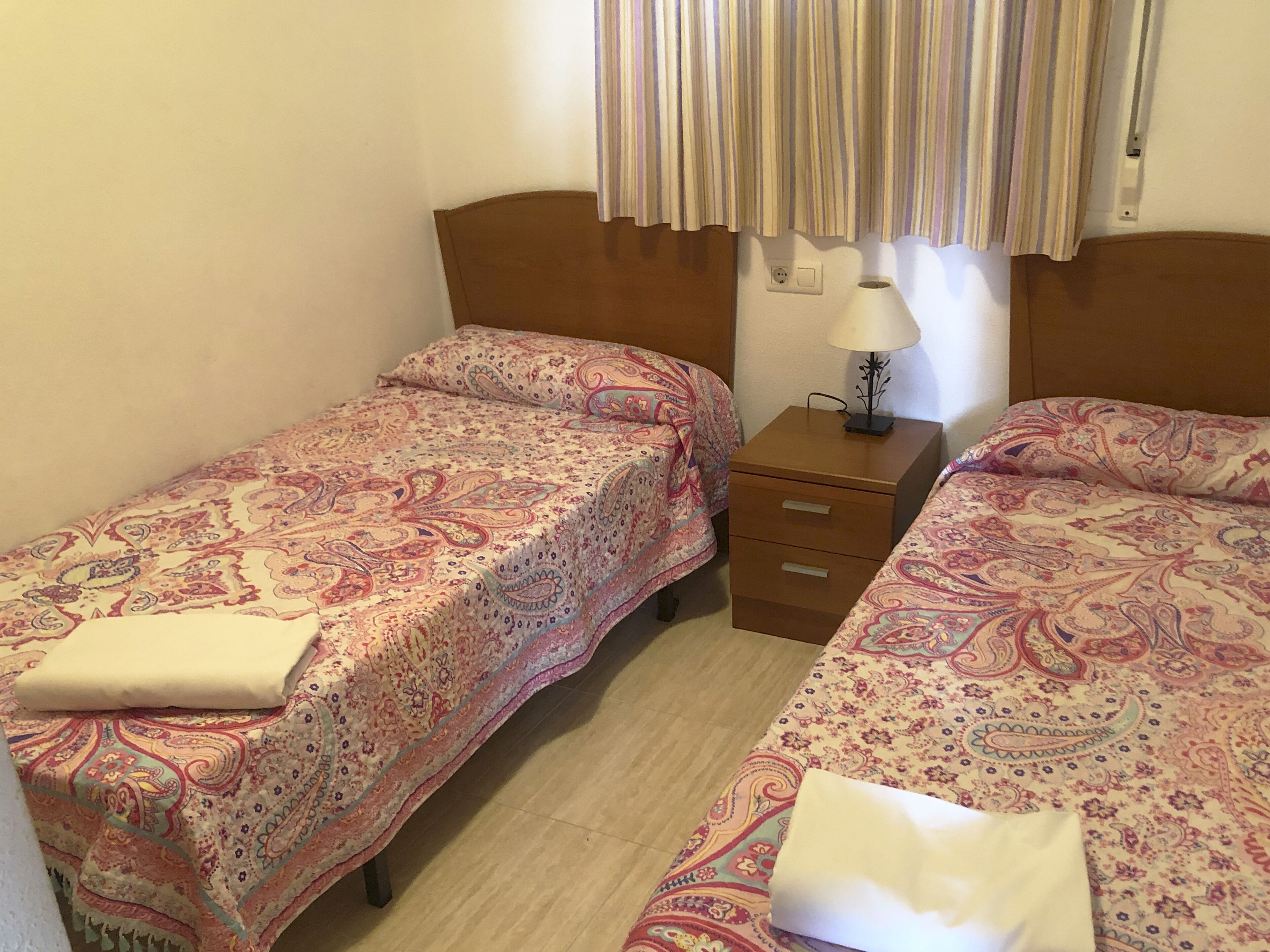 A small room with two beds, each with a patterned bedspread and a pillow. There is a nightstand with a lamp between the beds.