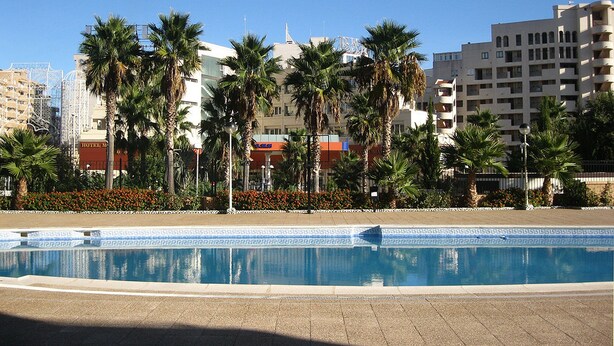 A swimming pool with a geometric tile pattern, surrounded by palm trees and a hotel building in the background. - Apartamentos Marina D'Or 3000 (Oropesa del Mar)