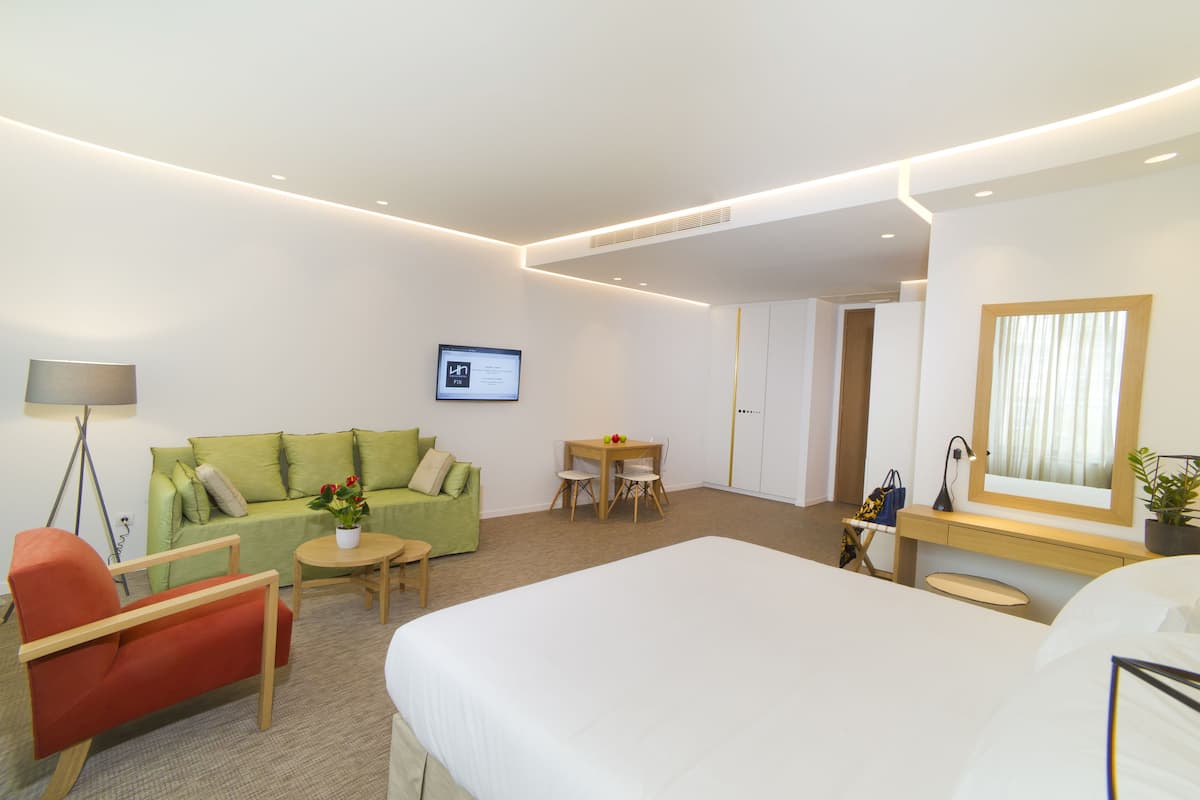 A hotel room with a bed, a green sofa, a red armchair, a small table with a lamp, and a desk with a computer monitor.