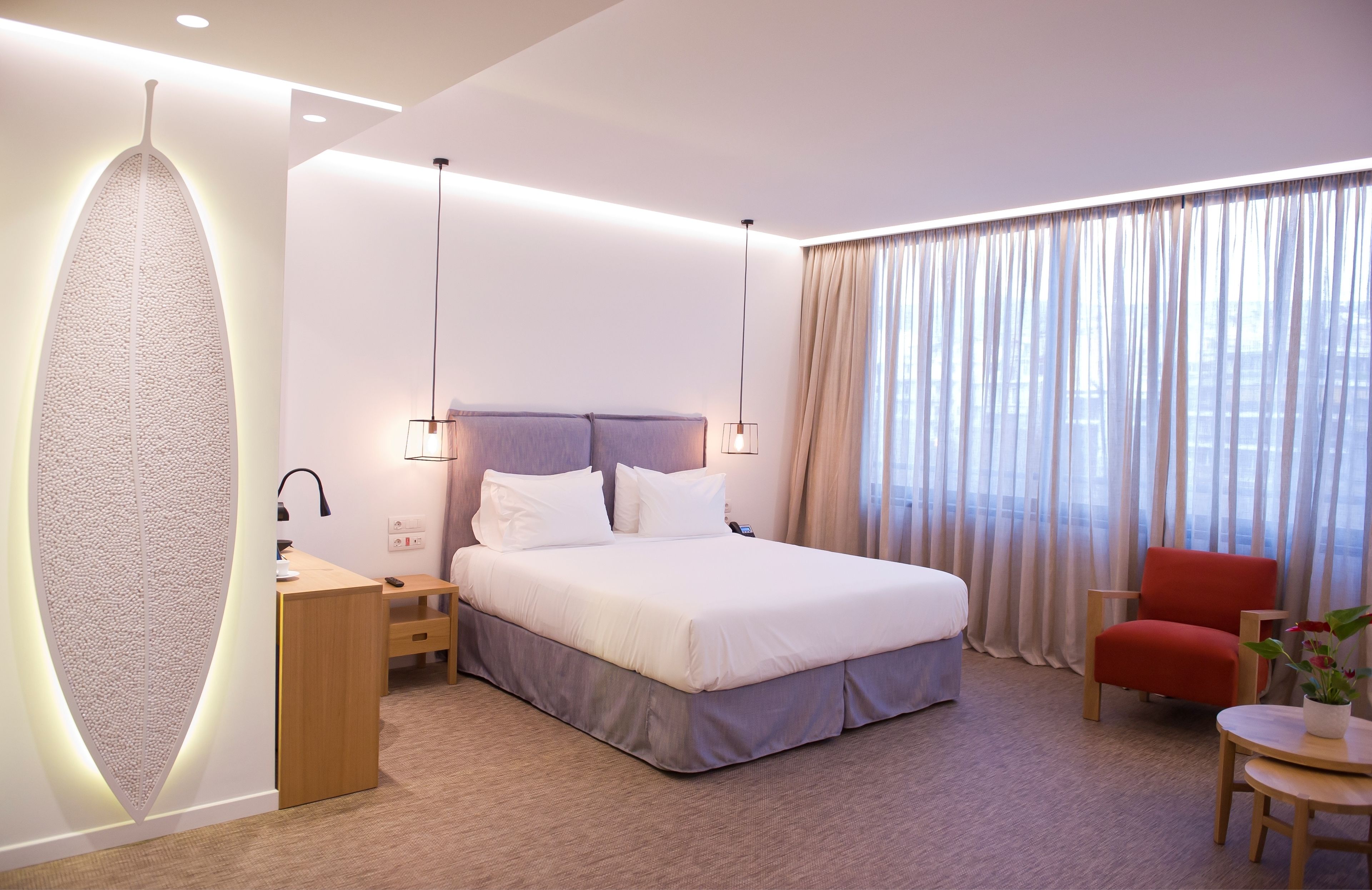 A modern hotel room with a large bed, bedside tables, a desk, a chair, and a large window with curtains.