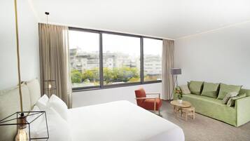 Una habitación de hotel moderna con un ventanal grande, una cama, un sofá verde, una mesita y una silla.