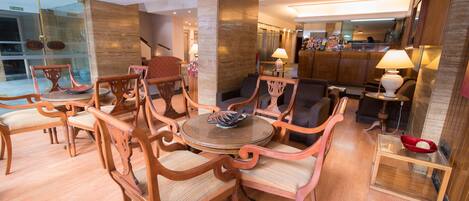 A spacious lobby with a round wooden table surrounded by ornate chairs, a glass partition, and a reception area in the background.