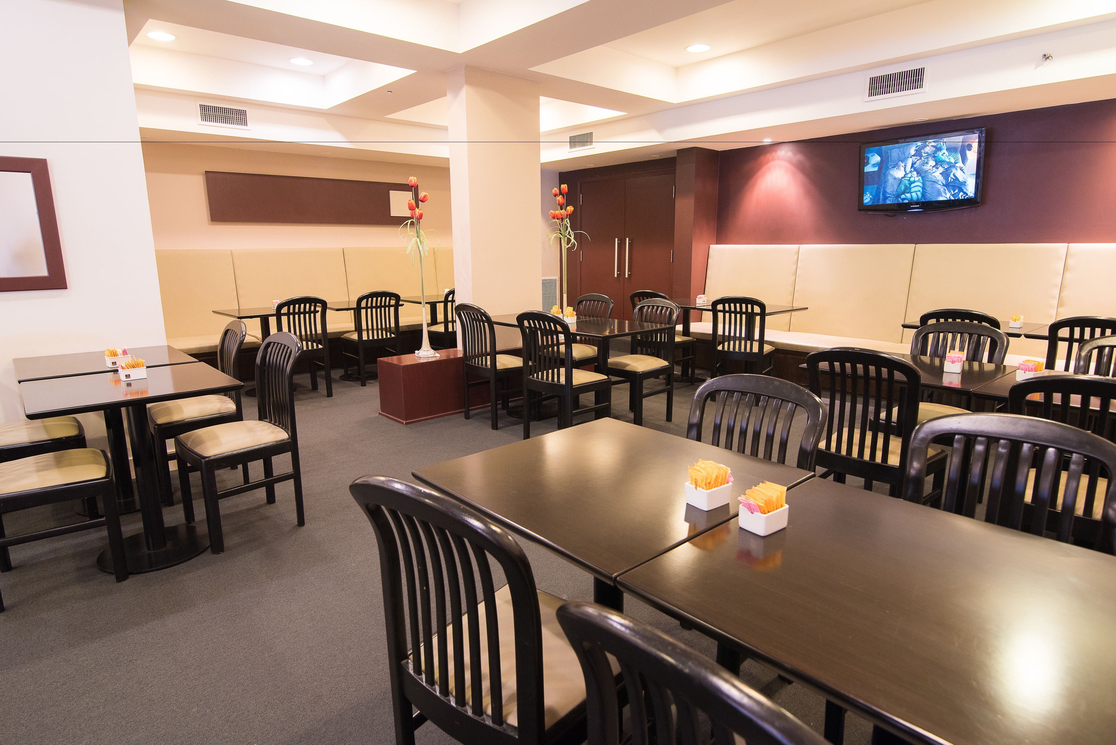 A hotel dining area with multiple tables and chairs, a television mounted on the wall, and a decorative vase with flowers.