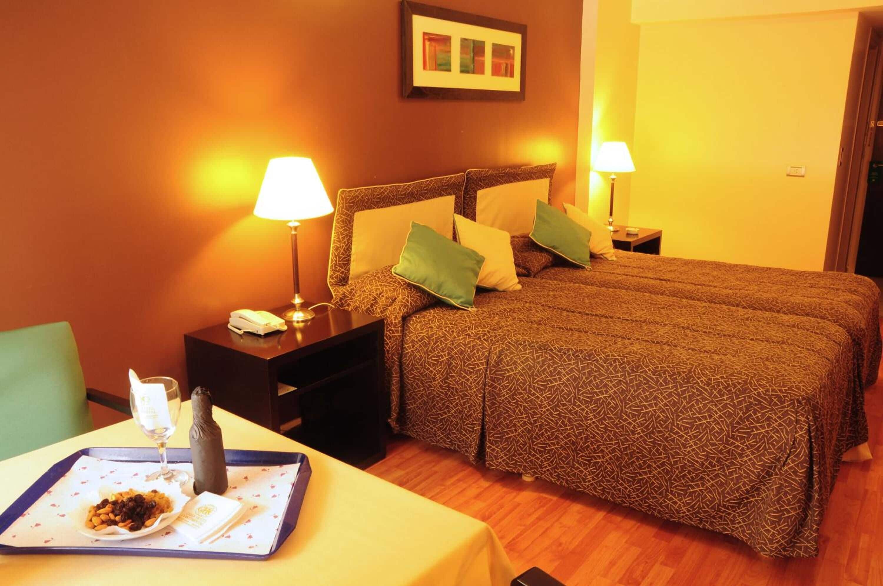 Quarto de hotel com uma cama grande, duas mesas de cabeceira com luminárias, uma pequena mesa com uma bandeja de comida e uma garrafa, e um quadro na parede.