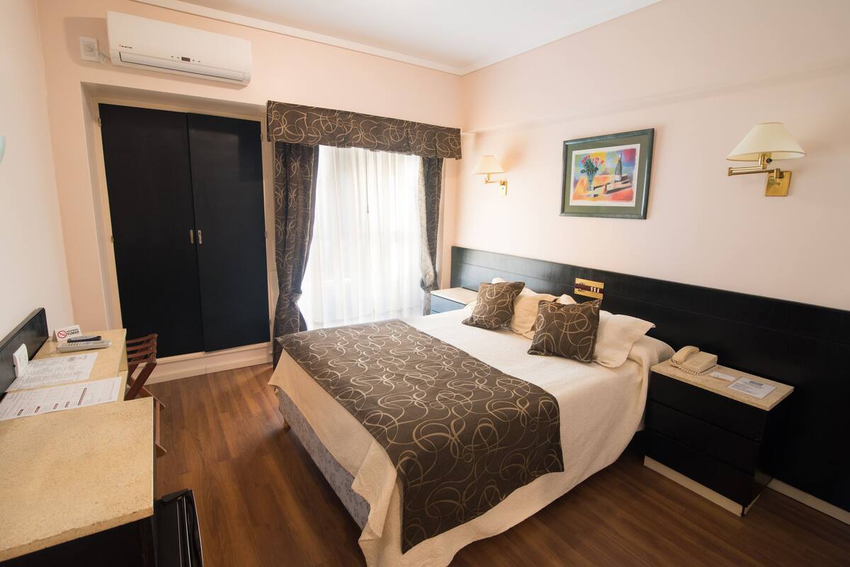 Una habitación de hotel con cama, mesita de noche, lámpara, una obra de arte y un escritorio con papeles.