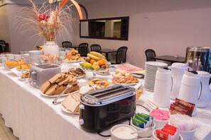 Une table de buffet proposant une variété d’aliments, notamment du pain, des fruits et des pâtisseries, ainsi que plusieurs distributeurs de café et de thé.