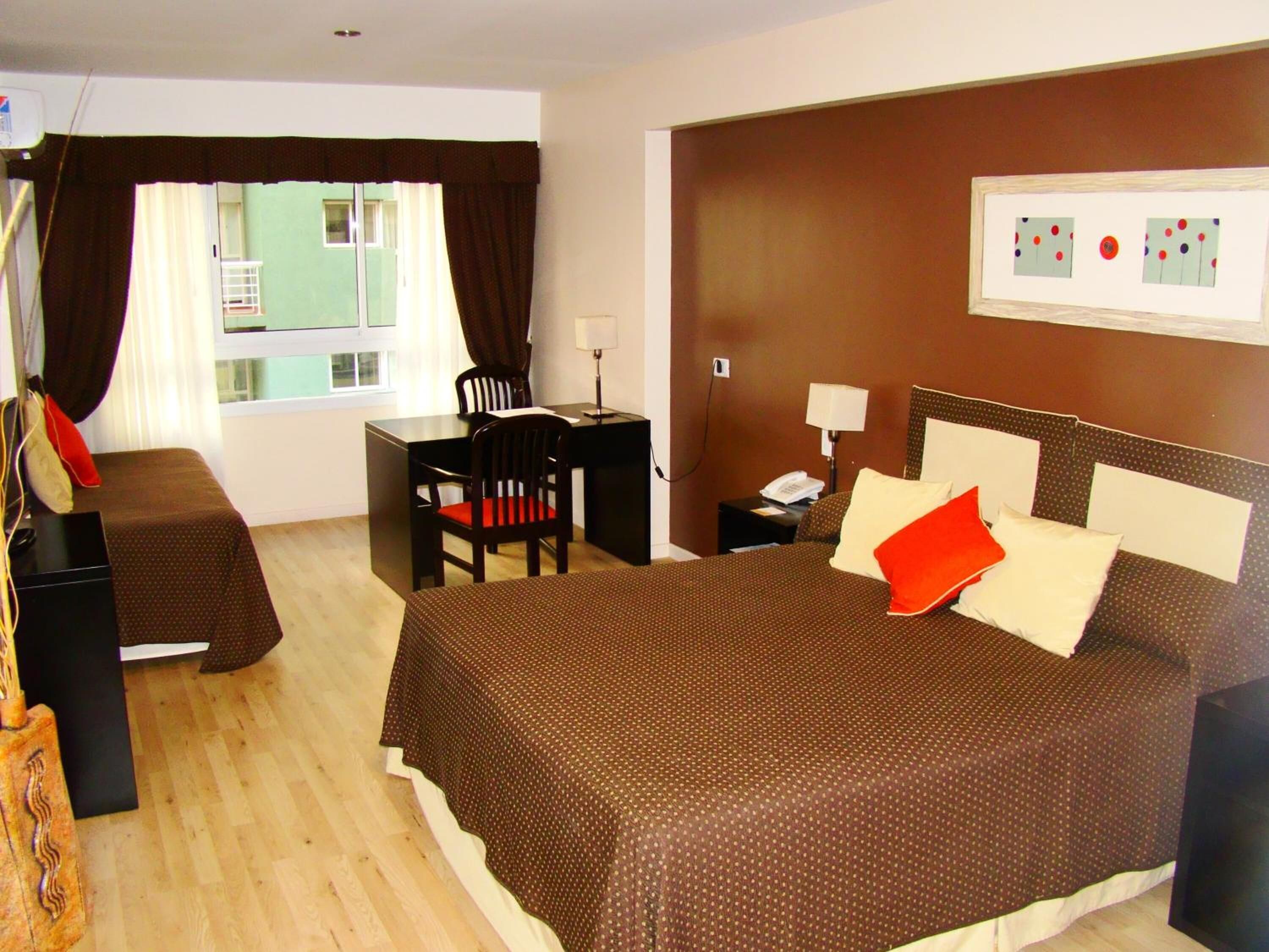 A hotel room with a large bed, a desk, a chair, a nightstand, and a window with curtains.