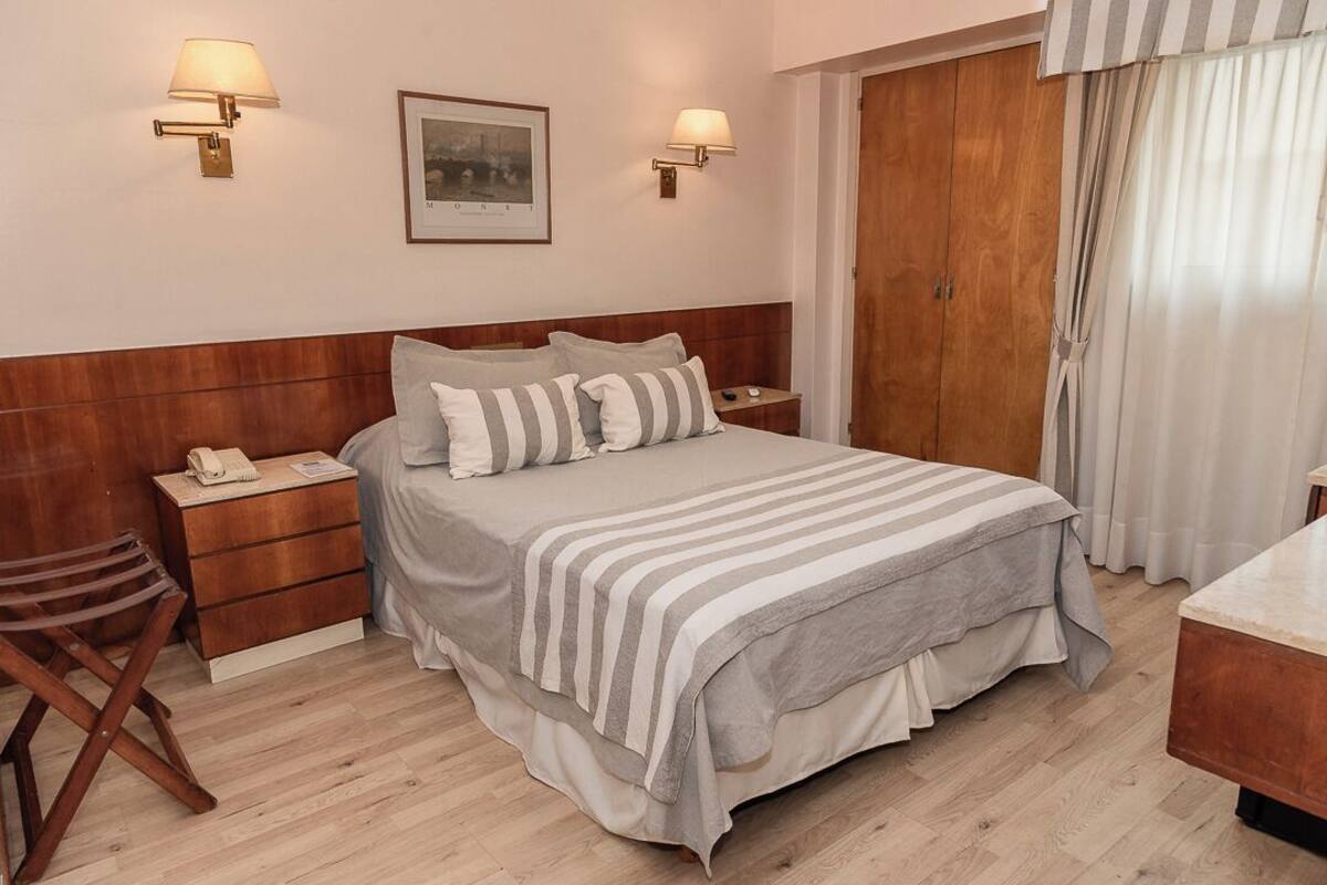 Un dormitorio ordenado con cabecera de madera, cama con ropa de cama a rayas, lámparas de noche, mesita de noche con un teléfono, una silla de madera y un armario.