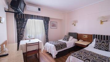 Habitación de hotel con dos camas individuales, una mesita con una silla, un televisor en la pared y una ventana con cortinas.