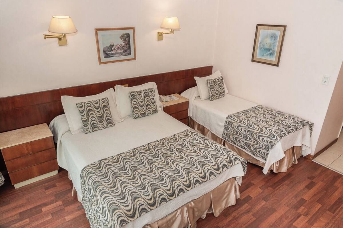 Una habitación de hotel con dos camas, cada una con ropa de cama estampada y cabeceros de madera. Hay lámparas de pared y cuadros enmarcados en las paredes.