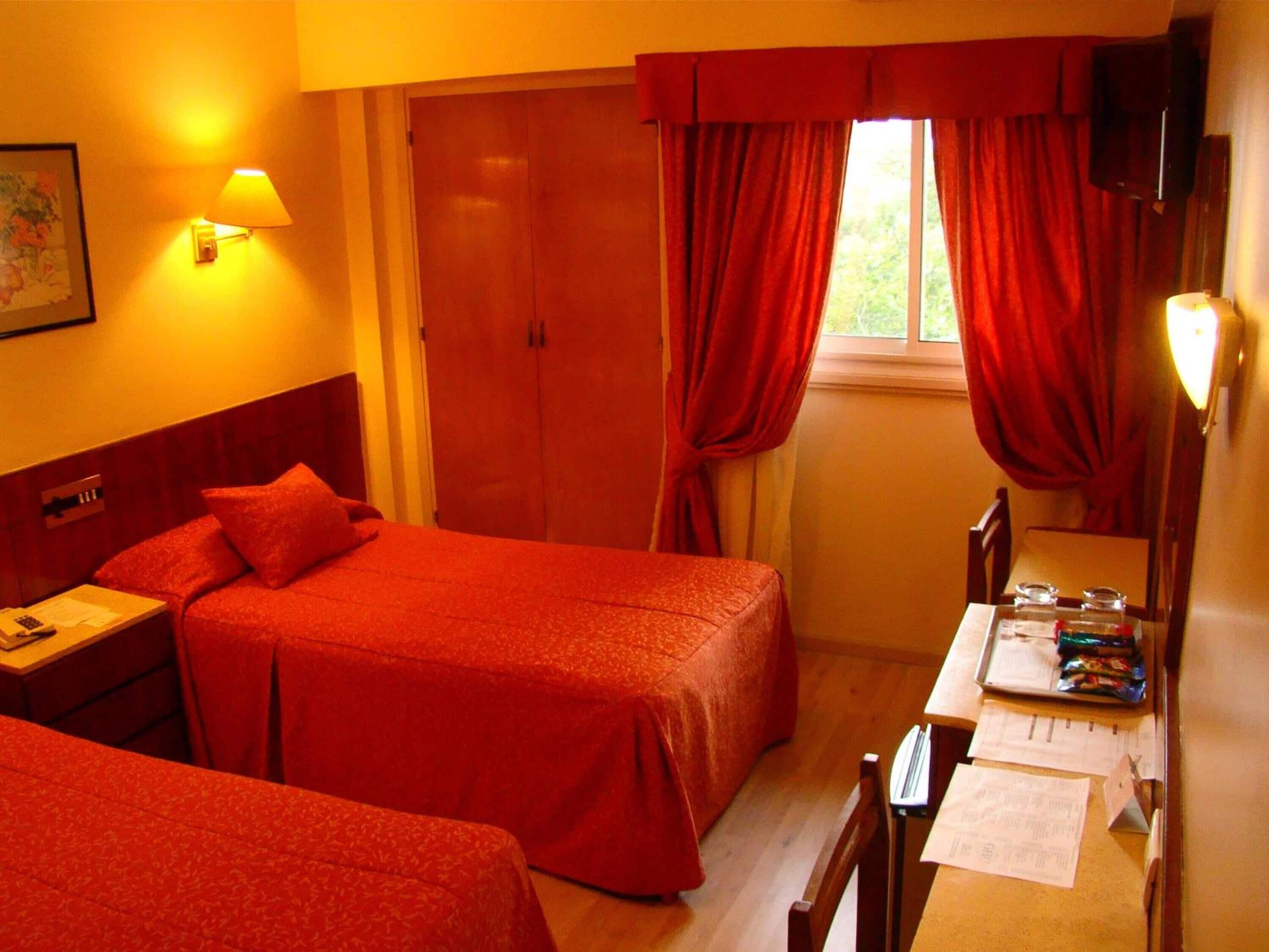 A hotel room with two beds, a desk with papers, a lamp, and a window with curtains.