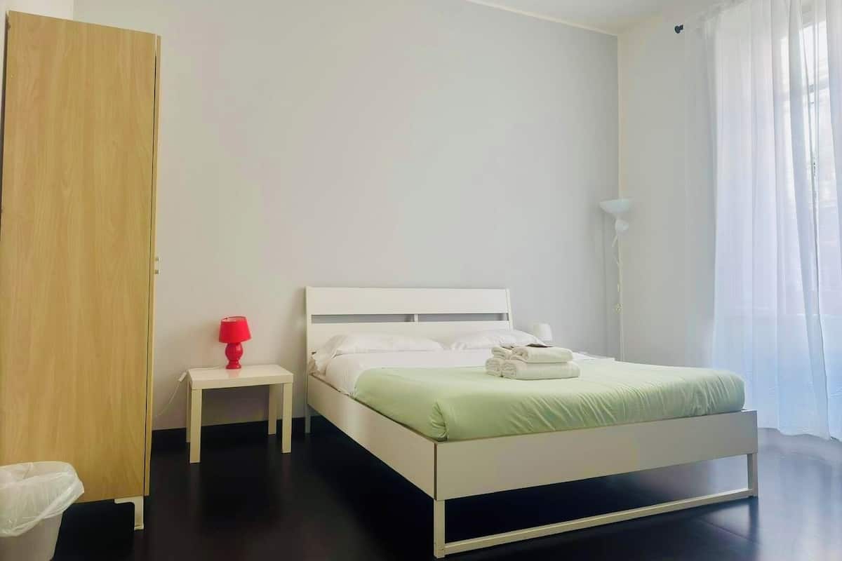 Un dormitorio minimalista con una cama blanca, una mesita de noche pequeña, una lámpara roja y un armario de madera.