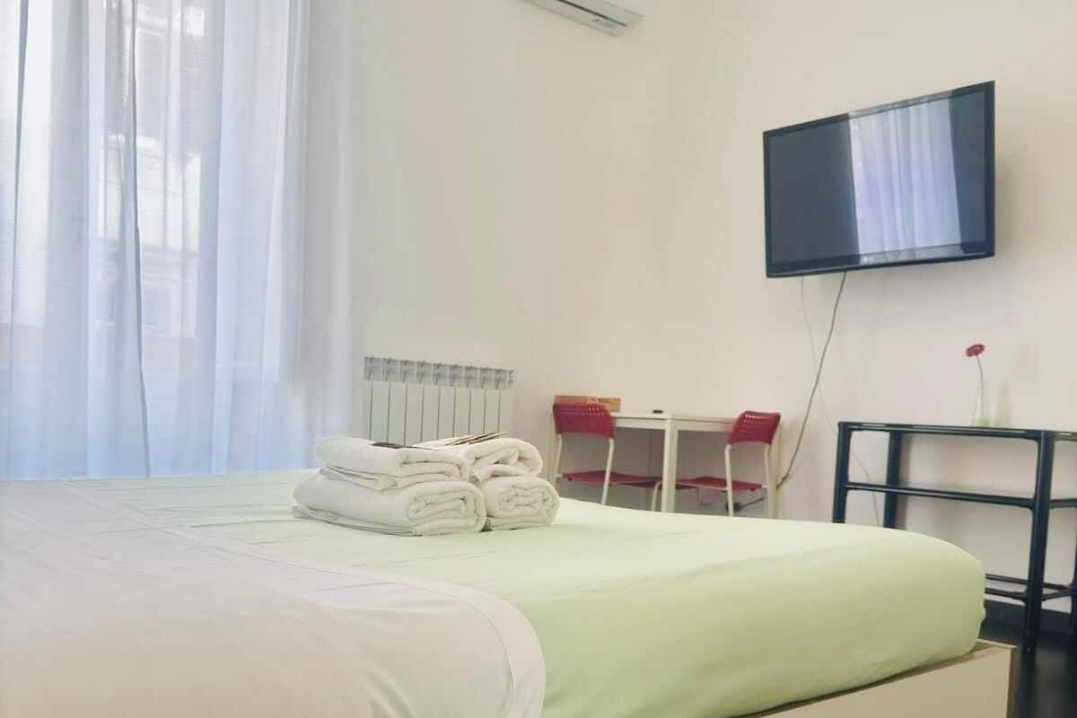 Una habitación de hotel con cama, televisor montado en la pared y ventana con cortinas traslúcidas.