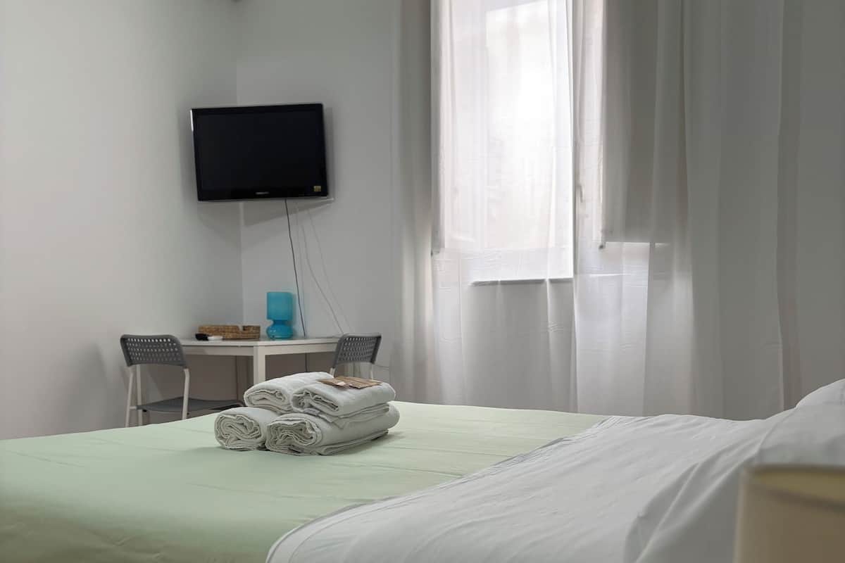 Habitación de hotel con cama, escritorio con silla, televisor de pantalla plana montado en la pared y ventana con cortinas traslúcidas.