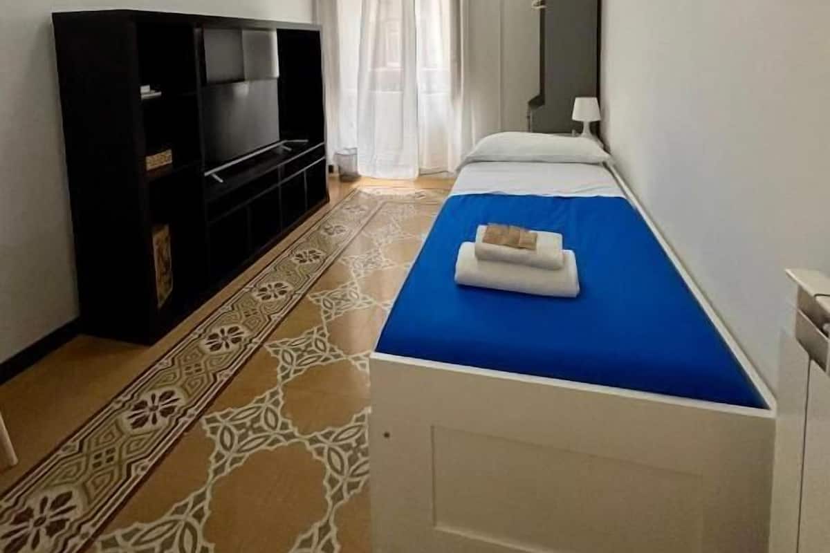 Una habitación pequeña y limpia con cama, televisión y ventana con cortinas.