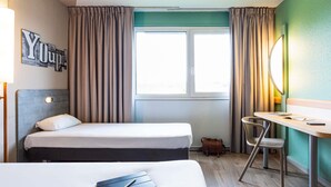 Habitación de hotel con dos camas, un escritorio, una silla, una mesita, una lámpara, un espejo y una ventana con cortinas.