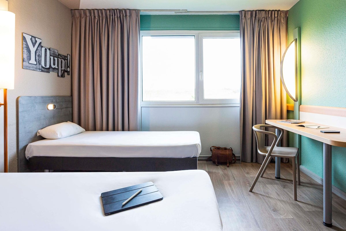 A hotel room with two beds, a desk, a chair, a small table, a lamp, a mirror, and a window with curtains.