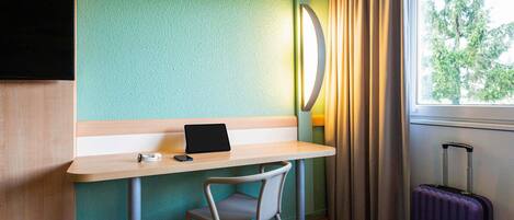 A hotel room with a wooden desk, a chair, a bed, a window with curtains, and a suitcase.