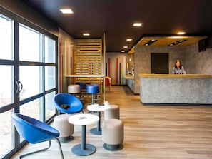 A modern interior with a reception desk, seating area with chairs and tables, and large windows. - ibis budget Angoulême Nord (Champniers)