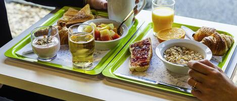 Bandeja de desayuno con tostadas, cereal, fruta, jugo y un croissant sobre una mesa junto a una ventana.