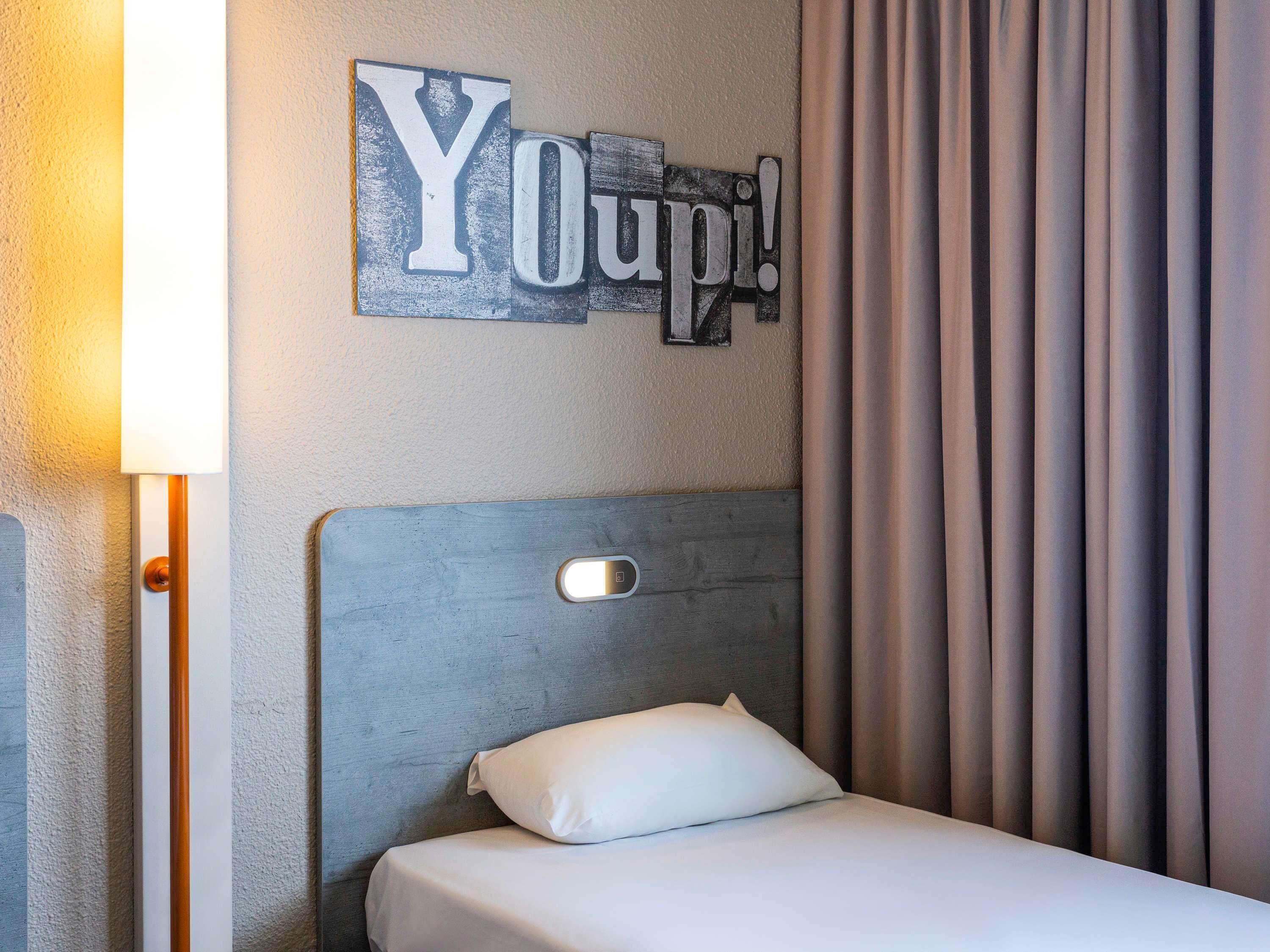 A hotel room with a bed, a headboard, a wall-mounted sign, a lamp, and curtains.