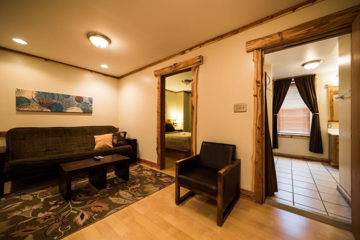 A living room with a sofa, a chair, a coffee table, and a rug. There is a painting on the wall and a wooden door frame.