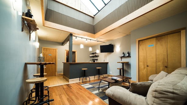Un vaste espace de vie doté d’un haut plafond, d’un plancher en bois, d’un canapé, d’une table basse et d’un coin bar avec des tabourets.