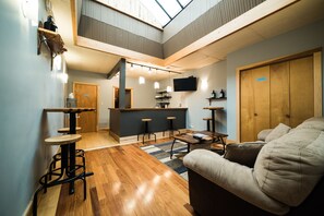 Un vaste espace de vie doté d’un haut plafond, d’un plancher en bois, d’un canapé, d’une table basse et d’un coin bar avec des tabourets.