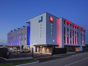 Un moderno edificio de hotel con letrero de 'ibis budget', entrada con portón y zona de estacionamiento.