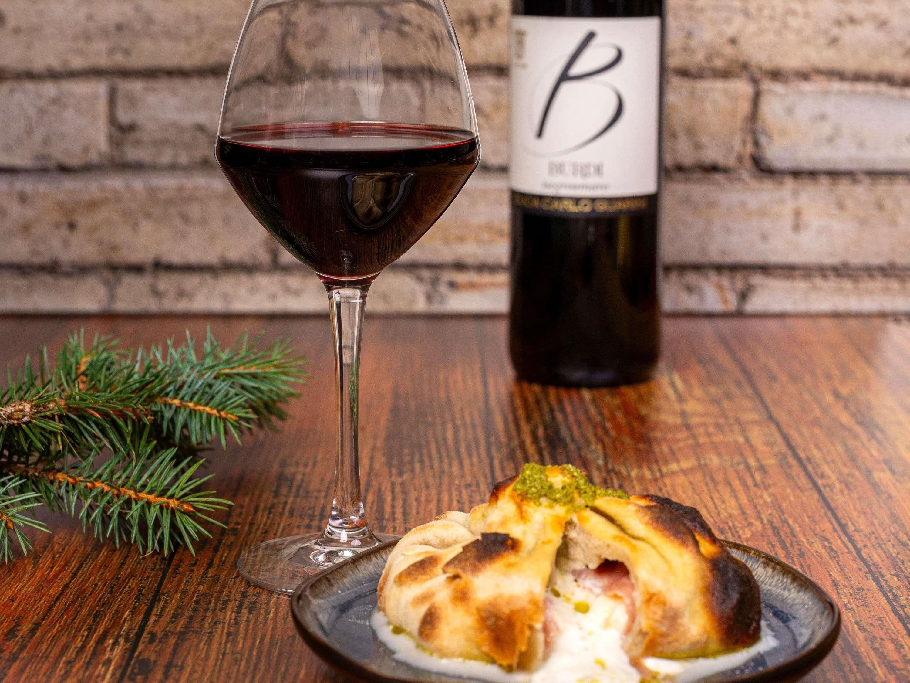 A glass of red wine, a bottle with a label, and a plate with food on a wooden table.
