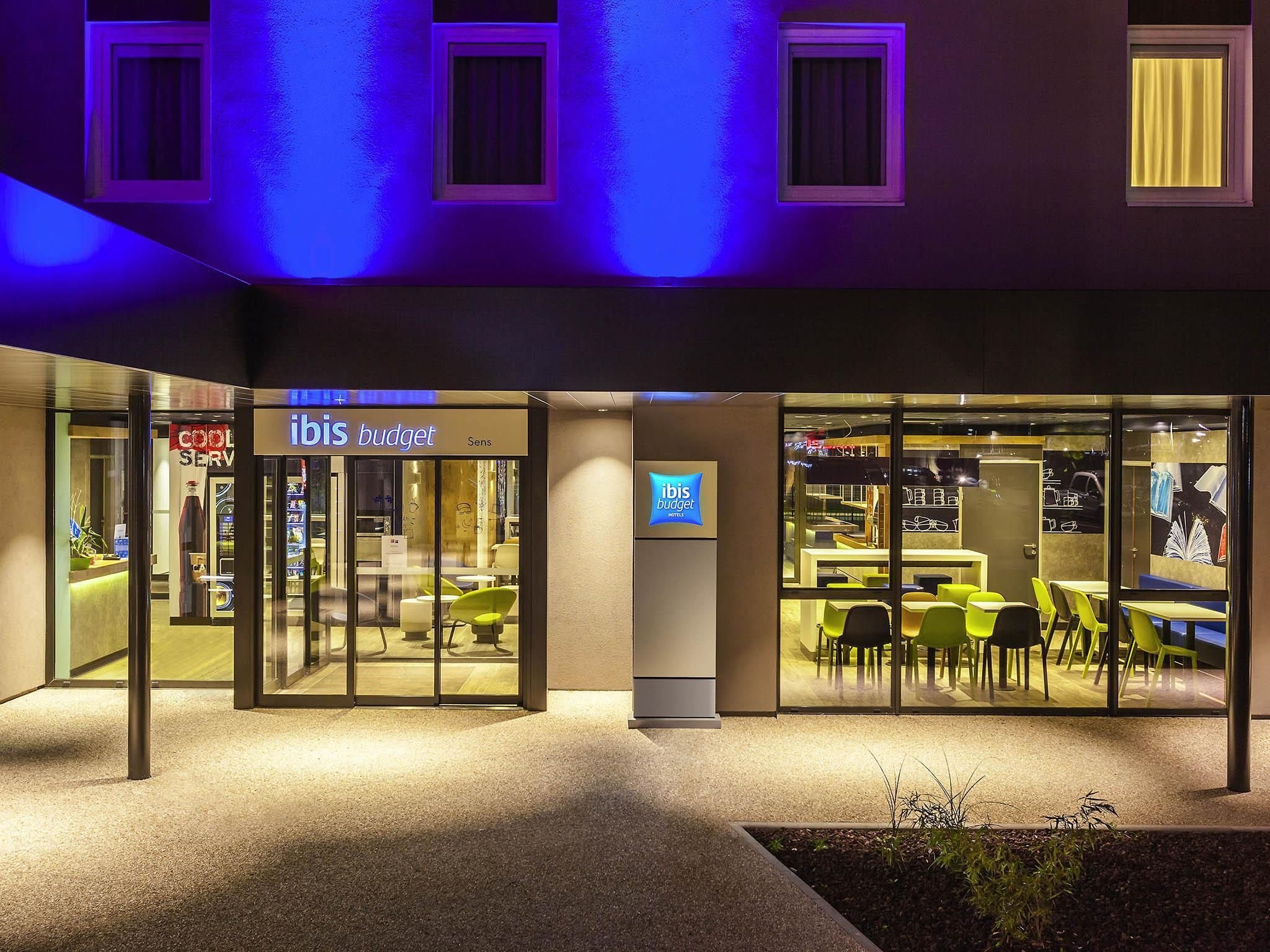 Un hotel moderno con fachada de vidrio, un letrero que dice 'ibis budget' y un área de estar visible a través de las ventanas.