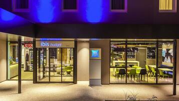 Un hotel moderno con fachada de vidrio, un letrero que dice 'ibis budget' y un área de estar visible a través de las ventanas.
