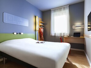 A hotel room with a bed, a desk with a laptop, a window with sheer curtains, and a wall-mounted light. - ibis budget Sens (Sens)