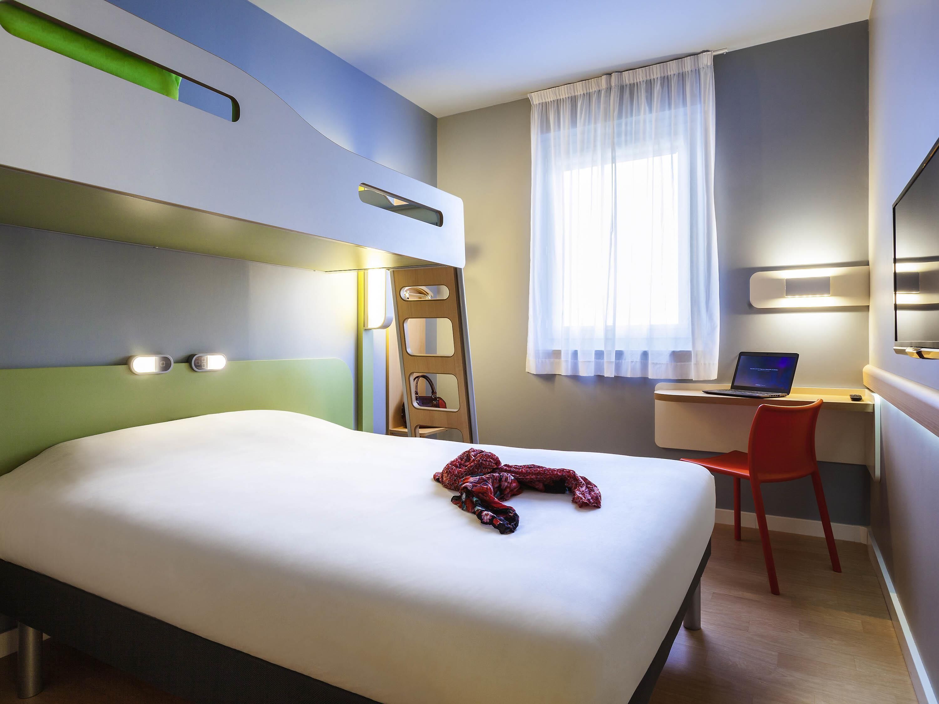 A hotel room with a bed, a desk with a laptop, a chair, a ladder, and a TV on the wall.