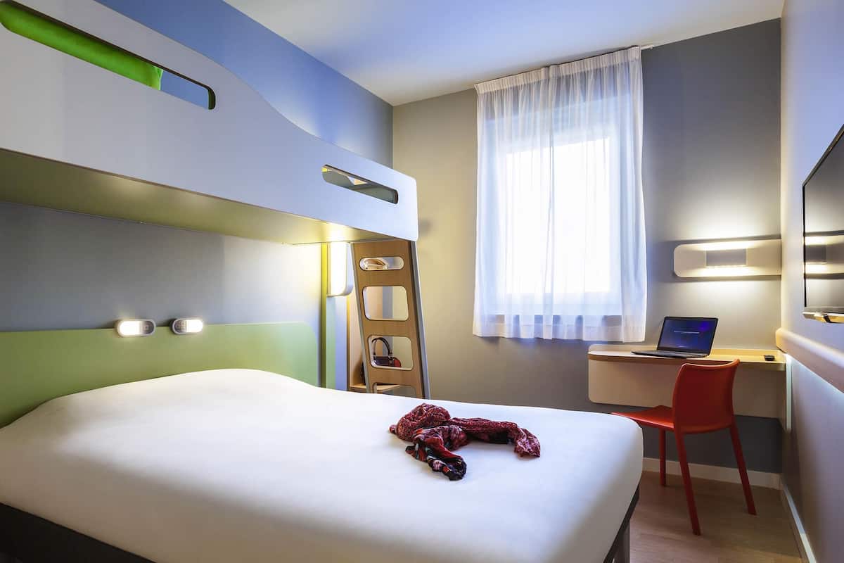 Quarto de hotel com cama, escrivaninha com laptop, cadeira, escada e televisão na parede.
