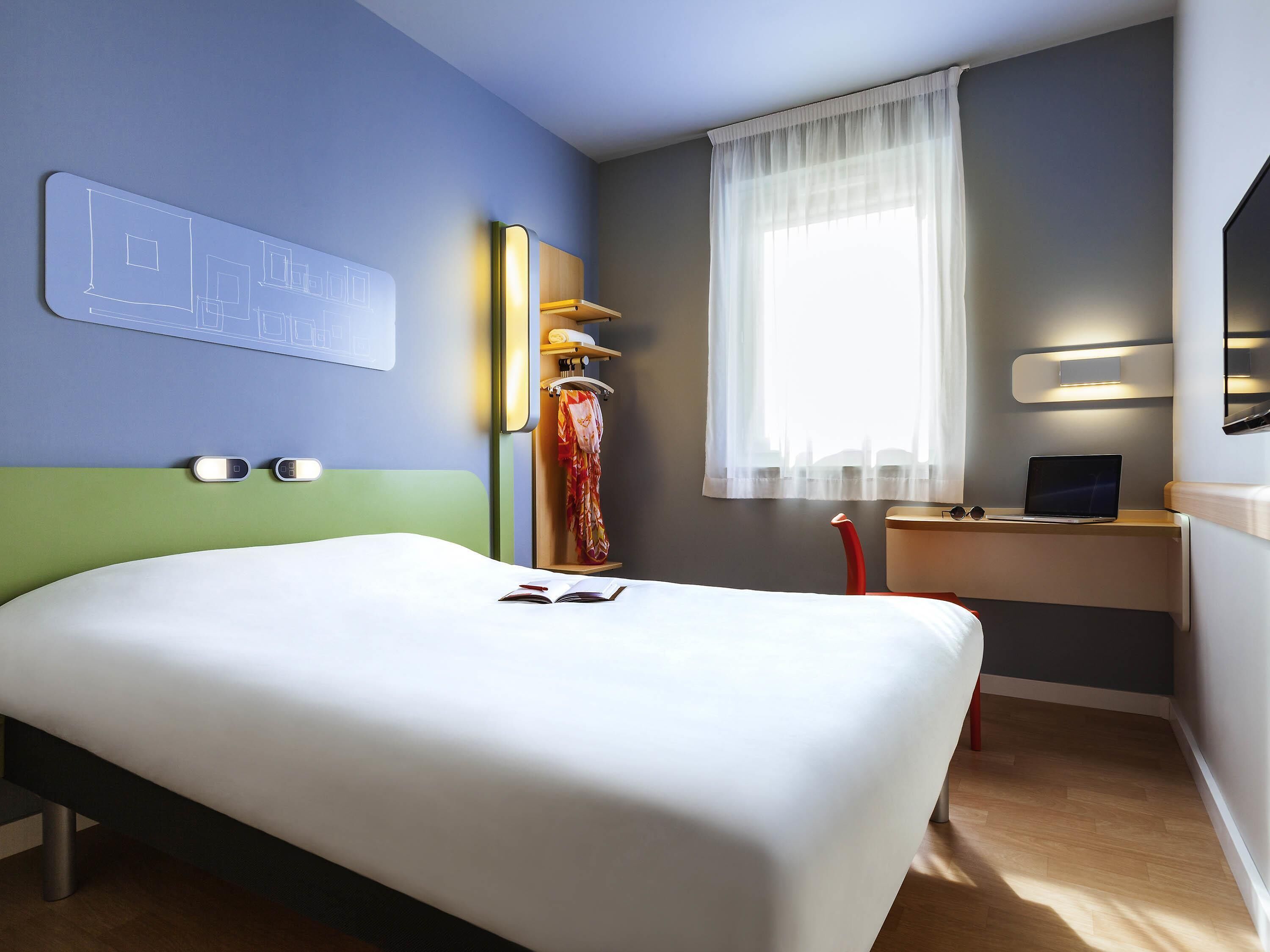 A hotel room with a bed, a desk with a laptop, a window with sheer curtains, and a wall-mounted light fixture.
