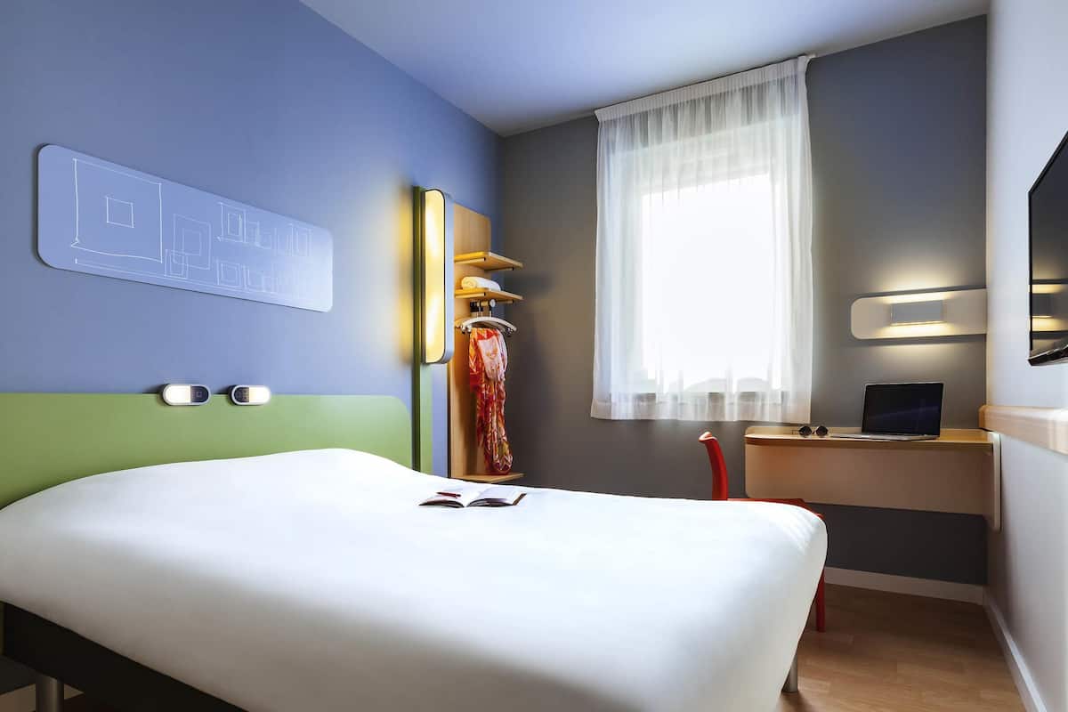 Quarto de hotel com cama, escrivaninha com laptop, janela com cortinas translúcidas e luminária fixada na parede.