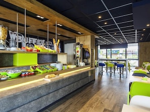 A modern café with a counter displaying food items, a coffee machine, and seating area with tables and chairs. - ibis budget Sens (Sens)