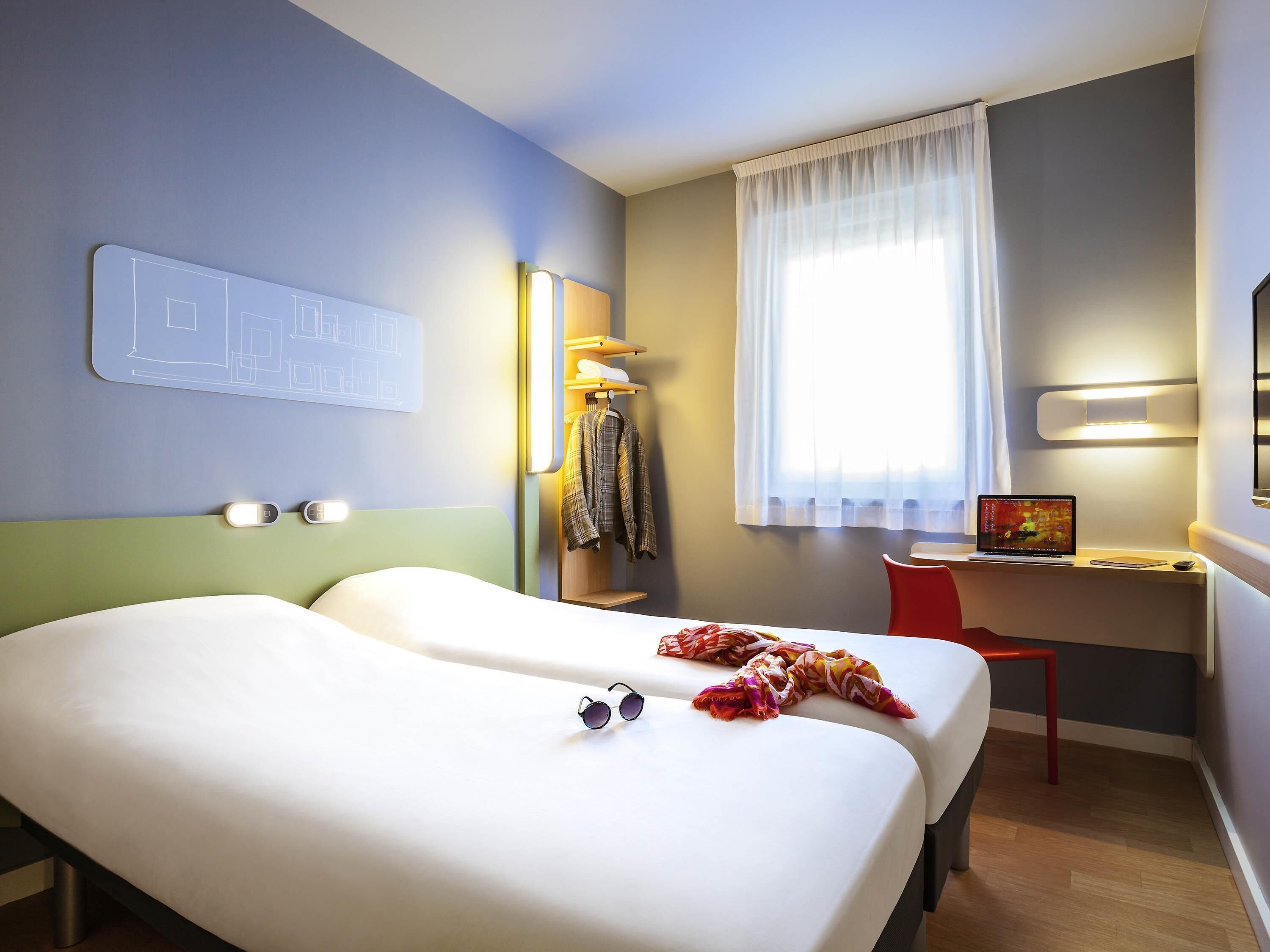 A hotel room with two beds, a desk with a laptop, a chair, a coat rack, and a window with curtains.