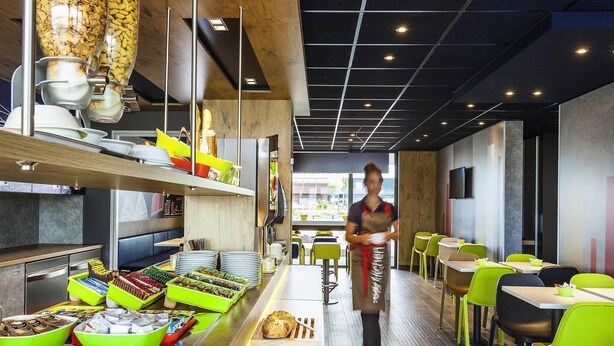 A modern restaurant with a long counter displaying various food items and a server walking down the aisle. - ibis budget Sens (Sens)