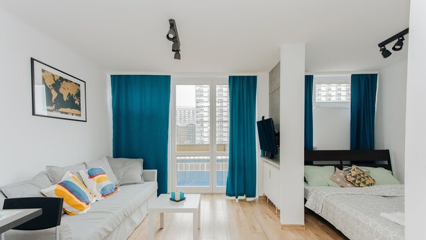 Et moderne, lysindtagende rum med sofa, lille bord, seng og et stort vindue med gardiner.