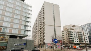 En høj, moderne bygning med flere etager og altaner, omgivet af andre bygninger og en gade med biler og vejskilte.