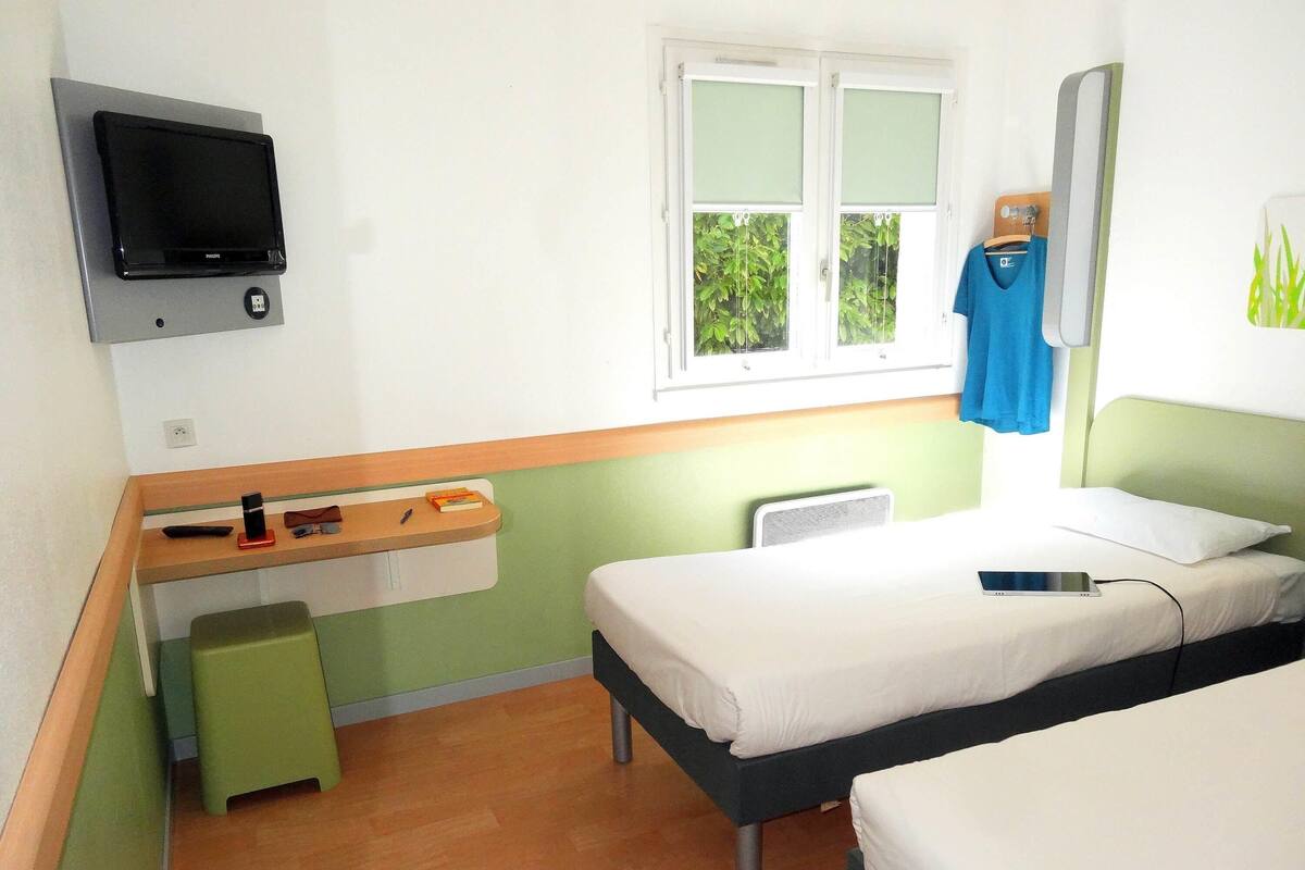 A small, simple room with two beds, a desk with a computer, a window with a view of greenery, and a wall-mounted TV.