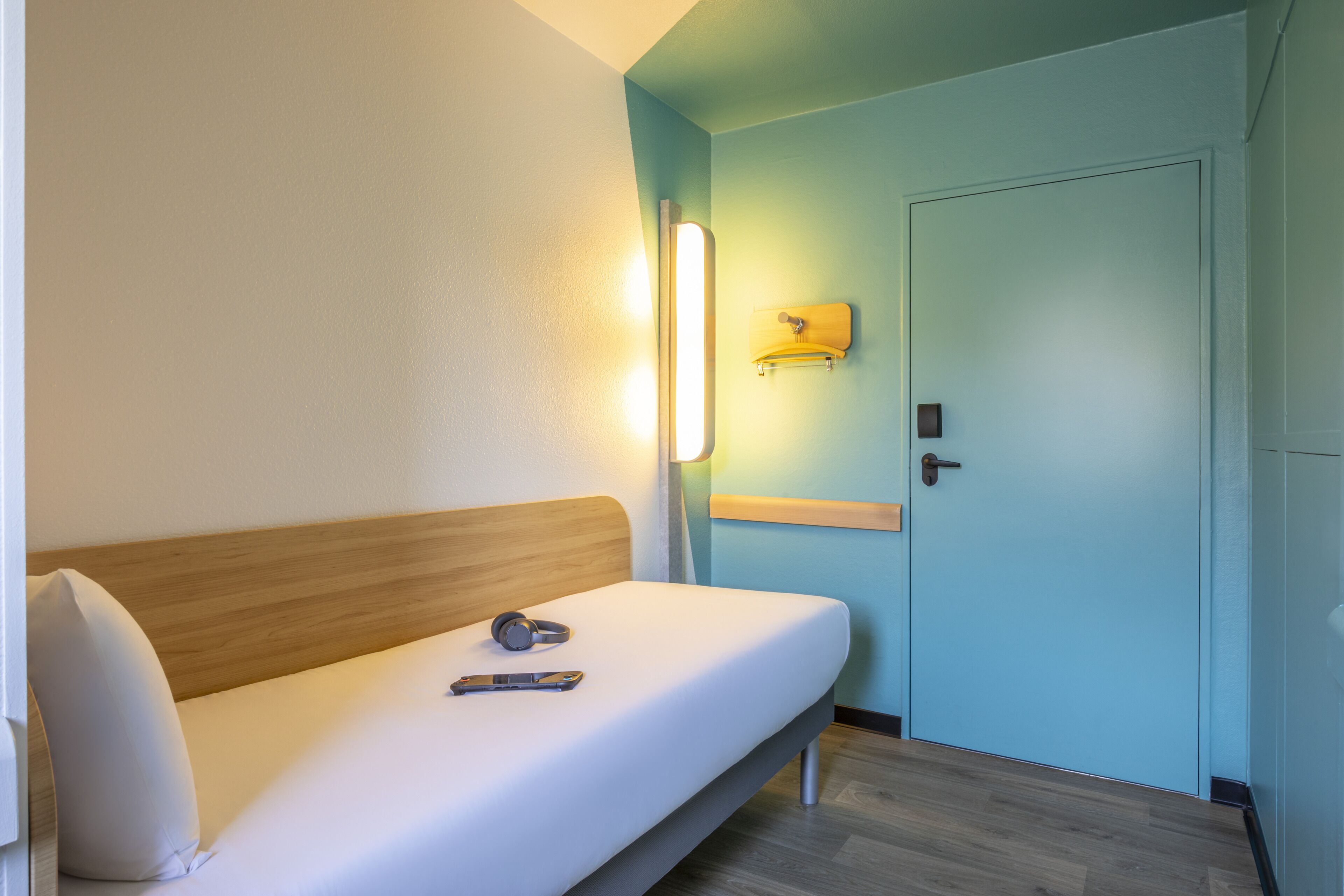 Una habitación de hotel con una cama individual, una mesita de noche, una lámpara de pared y una puerta.