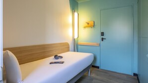 Quarto de hotel com uma cama de solteiro, mesa de cabeceira, luminária na parede e uma porta.