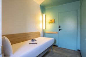 Une chambre d’hôtel avec un lit simple, une table de chevet, une lampe fixée au mur et une porte.