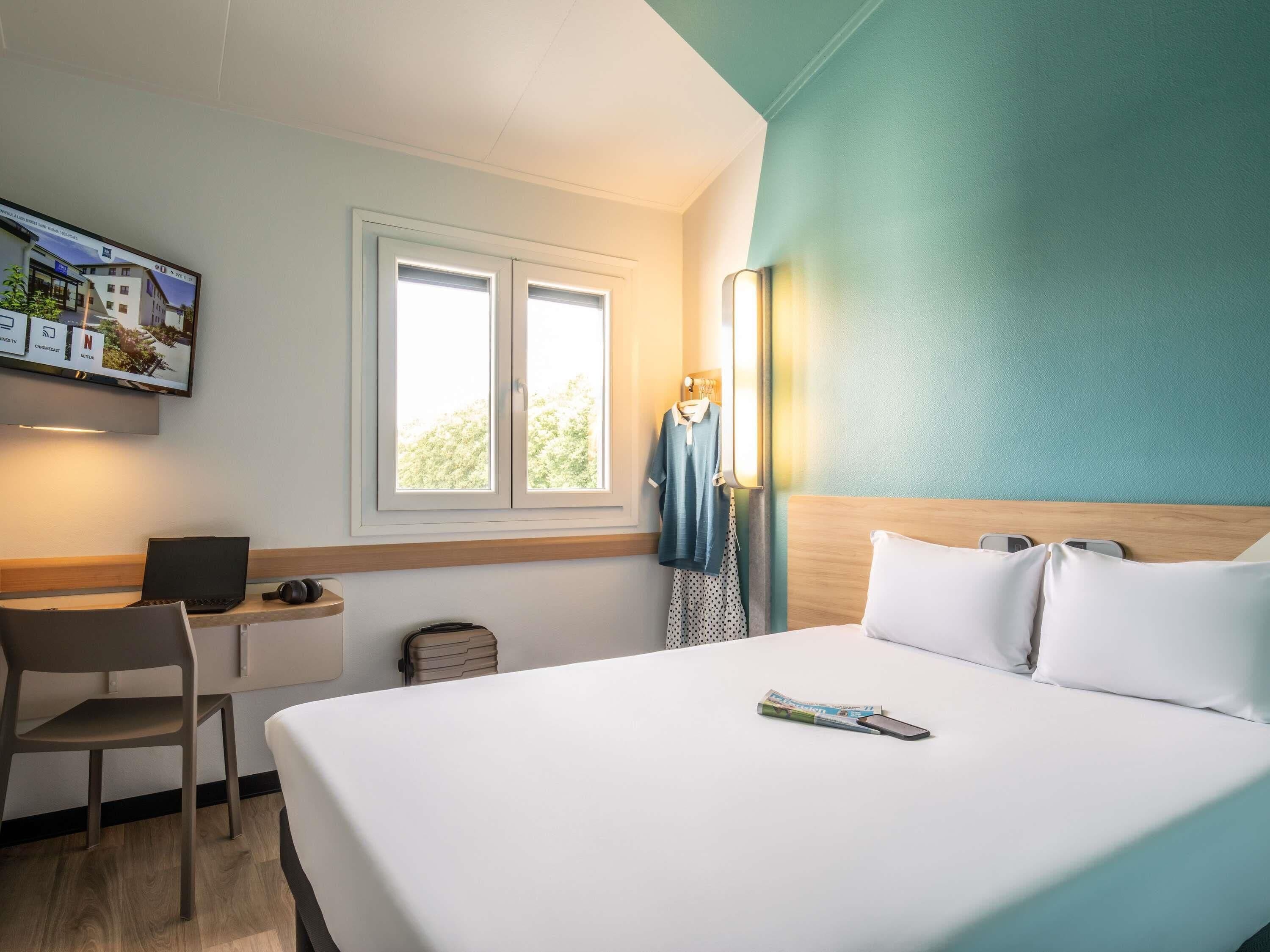 Habitación de hotel con una cama, un escritorio con una computadora portátil, una silla, una maleta y un televisor montado en la pared.